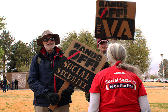 Robert Angelo and Robert Scott stand with signs that read “Hands Off! Social Security” and “Hands off! The VA,” and Joan Crowley wears a shirt that reads “Social security is on the line.”