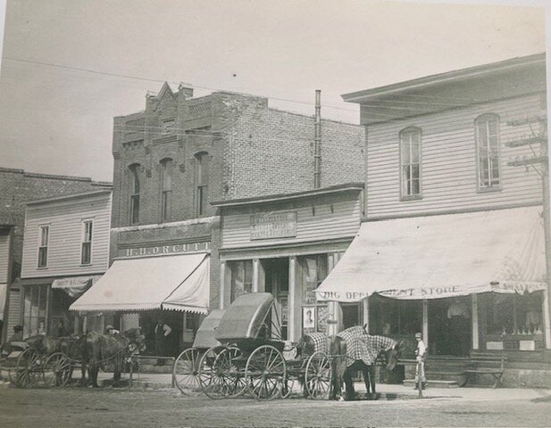 Quackenbush Music Store, short building second from the right.  The sign reads D.Quackenbush,          David Quackenbush, in the middle with his fiddle, surrounded by A.O.U.W. members Musical Instruments and Strings